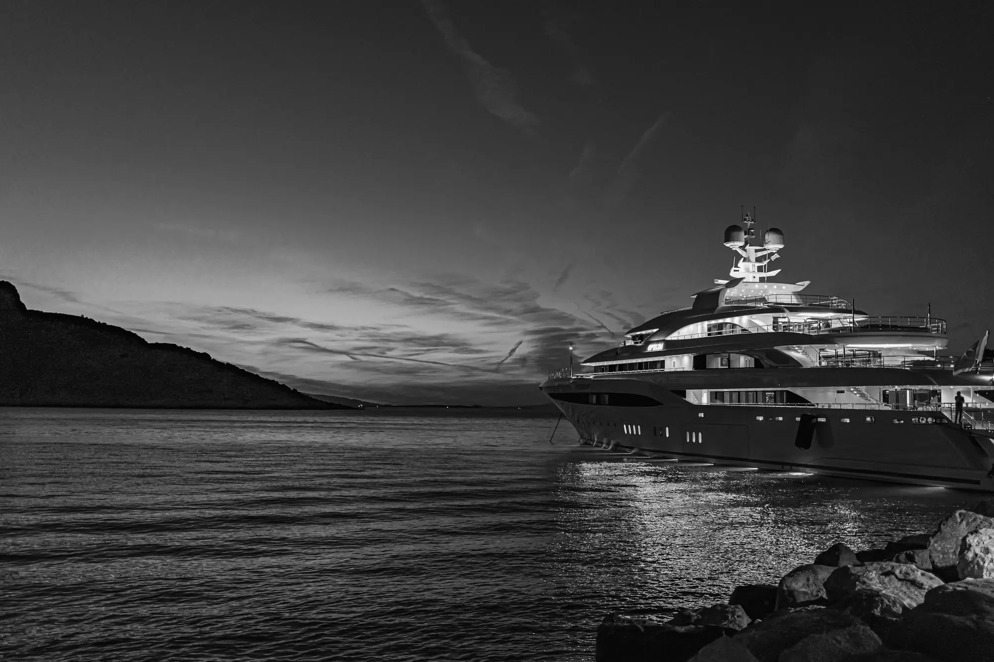 A megayacht at anchor in a bay at dusk, lit along every deck